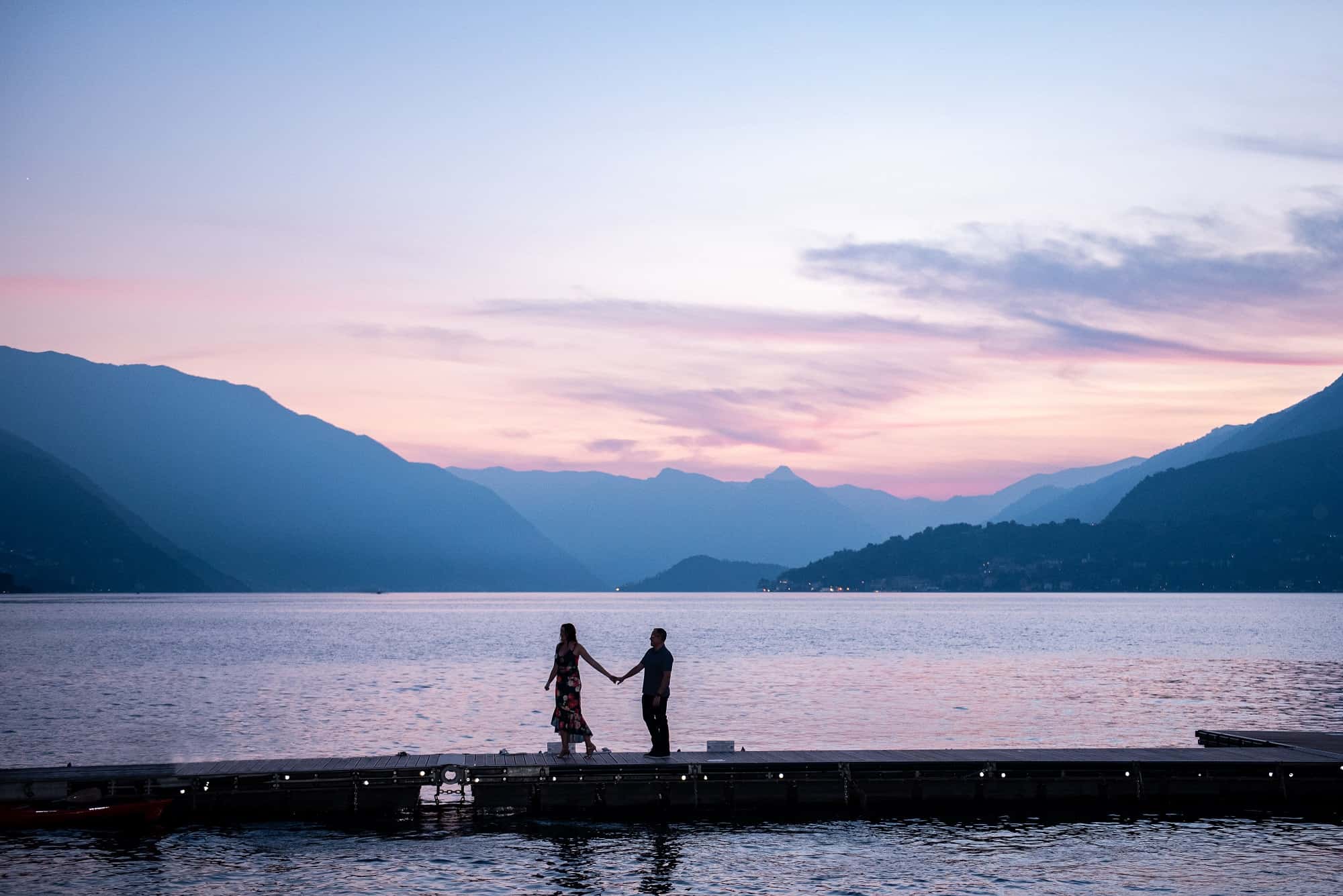 sunset with the italian mountains in the background and the couple on the lake holding hands, walking silhouetted