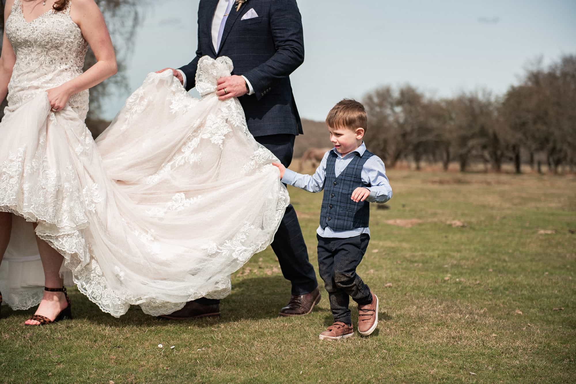 Groom holding brides dress on their wedding day with their little boy helping hold the dress